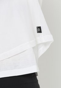 Vit lagerpålägg med ett genomskinligt ytterlager och ett solitt vitt innerlager. Har en svart etikett med en logotyp på nederkanten.