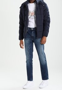 Námořnicky modrá prošívaná bunda s kapucí, bílé tričko s grafikou, světle modré džíny a bílé tenisky. Bunda má prošívanou texturu a zipové zapínání.