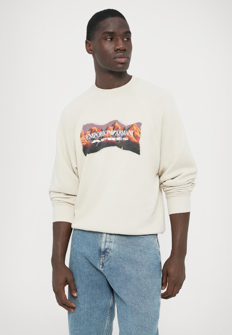 Light beige sweatshirt with a textured finish, featuring a multicolored graphic of mountains and text on the front. Pair of blue denim jeans.