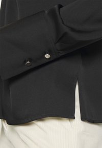Maglietta nera a maniche lunghe in tessuto con una texture liscia, caratterizzata da dettagli con bottoni argentati sui polsini e uno spacco laterale.