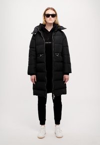 Cappotto gonfiabile nero con chiusura a zip, due dettagli a toggles e cappuccio staccabile, indossato sopra un hoodie nero e pantaloni, abbinato a sneaker bianche.