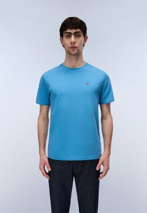 Junger Mann mit dunklen Haaren, der ein schlichtes blaues T-Shirt und dunkle Jeans trägt, vor einem hellgrauen Hintergrund stehend.