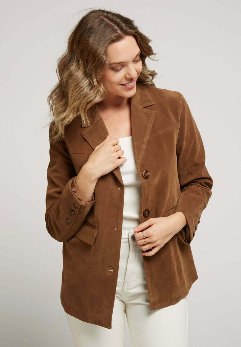 Blazer en daim marron avec revers en notch et fermeture à bouton, doté de manches longues, de poches latérales et d'une texture lisse. Porté par-dessus un haut blanc.