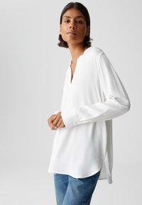 Vrouw met kort donker haar, gekleed in een losse witte blouse met lange mouwen en blauwe jeans, staand tegen een effen lichtgrijze achtergrond.