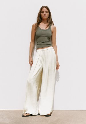 Une femme est debout contre un mur blanc, portant un haut sans manches vert olive, un pantalon blanc ample à jambes larges et des sandales plates noires.