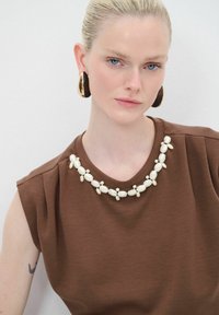 Top sin mangas marrón con un cuello decorativo de cuentas blancas. La modelo lleva pendientes de aro dorados brillantes. Tejido suave, diseño sencillo.