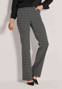 Vrouw in zwart-witte geometrisch patroon flare broek, zwarte puntige hakken en een zwarte top met één hand in de zak.