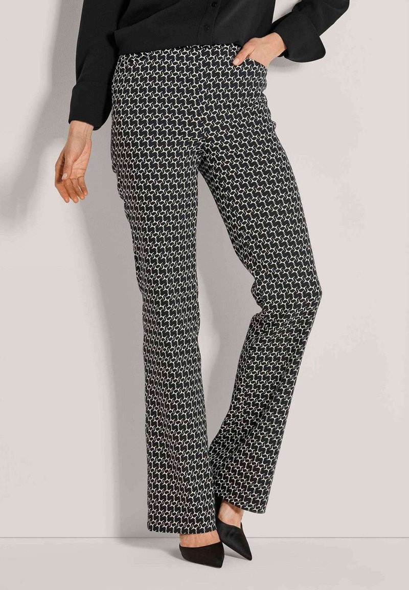 Vrouw in zwart-witte geometrisch patroon flare broek, zwarte puntige hakken en een zwarte top met één hand in de zak.