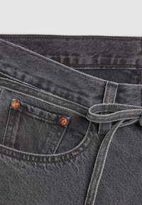 Gros plan d'un jean en denim gris montrant une poche avant avec des rivets en cuivre et une ceinture en tissu passée dans les passants.