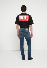 Schwarzes T-Shirt mit kurzen Ärmeln und rotem "DIESEL"-Logo auf dem Rücken, kombiniert mit blauen Slim-Fit-Jeans und schwarzen Turnschuhen, Ansicht von hinten.