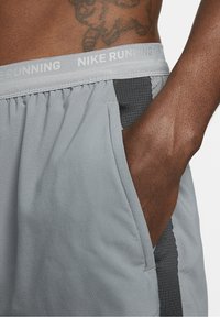 Homme portant un short de course Nike gris clair avec une main dans la poche, montrant une taille élastique et un panneau latéral gris foncé.