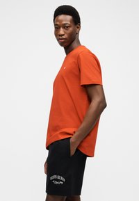 Νεαρός άνδρας με πορτοκαλί μπλουζάκι και μαύρα σορτς Calvin Klein, με το ένα χέρι στην τσέπη, στέκεται μπροστά σε απλό ανοιχτόφωνο φόντο.