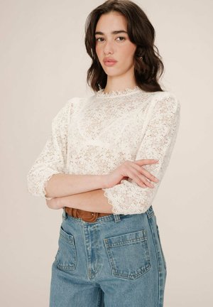 Blouse en dentelle blanche à manches trois-quarts, décolleté festonné et motifs floraux complexes, associée à un jean en denim bleu clair taille haute.