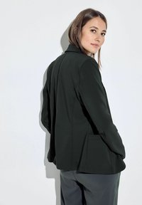 Blazer tailored di colore verde scuro con un design strutturato, caratterizzato da rever a punta, tasche frontali e una texture liscia, indossato sopra pantaloni grigi.