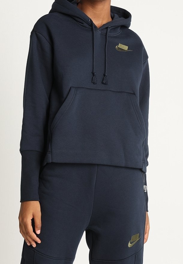 Persona que lleva una sudadera con capucha de Nike en azul marino con un bolsillo frontal y pantalones de chándal a juego, ambos con logotipos de la marca Nike en color verde oliva.