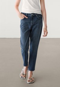 Donkerblauwe denim jeans met een relaxed fit, vijf zakken en een ritssluiting. Draag ze met gestreepte teenslippers en een getailleerd wit shirt.