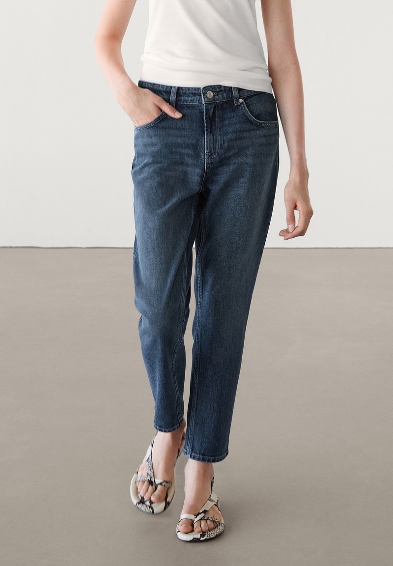 Donkerblauwe denim jeans met een relaxed fit, vijf zakken en een ritssluiting. Draag ze met gestreepte teenslippers en een getailleerd wit shirt.
