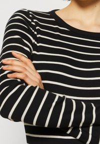 Maglietta nera a maniche lunghe con righe orizzontali bianche, caratterizzata da una texture a coste e collo a giro. Braccia incrociate davanti.