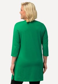 Grünes, langärmliges Tunika-Oberteil aus weichem Material, mit geradem Schnitt und seitlichen Schlitzen für Bewegungsfreiheit, getragen über schwarzen Leggings.