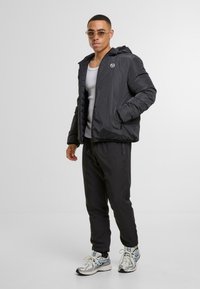 Chaqueta de plumas negra con capucha, cierre de cremallera y logo en el pecho. Combinada con pantalones jogger negros y zapatillas plateadas metálicas. Textura suave.