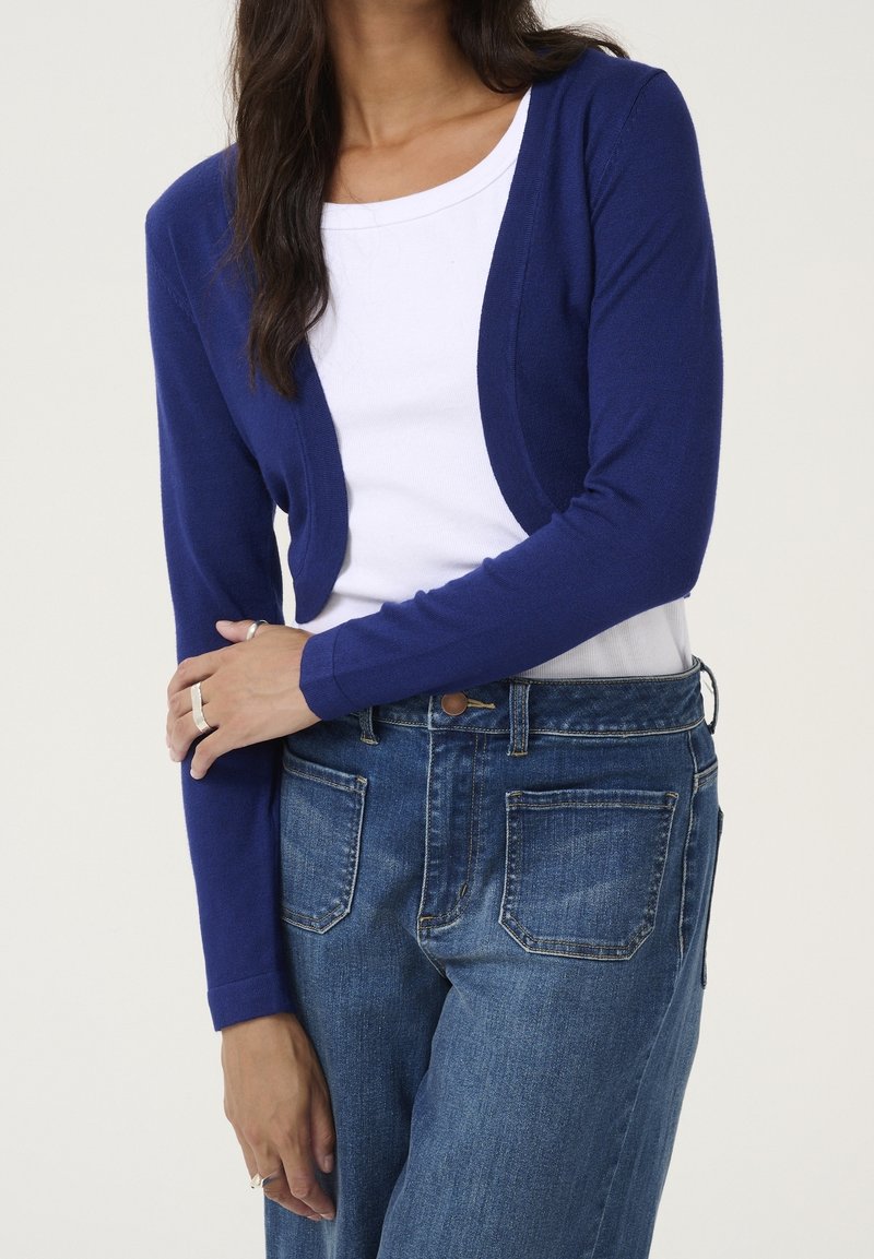 Femme portant un cardigan court bleu marine sur un top blanc et un jean bleu taille haute avec des poches plaquées à l'avant.