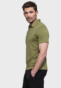 Grünes Poloshirt mit strukturierter Oberfläche, dreiknöpfigem Kragen und Logodetail am linken Ärmel, kombiniert mit schwarzen Hosen.