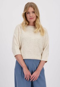 Crème kleurige geribbelde trui met driekwart mouwen, met een zachte textuur. Het model draagt lichtblauwe broeken, met de handen voor zich geklapt.