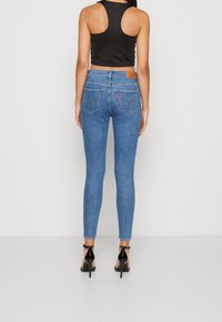 Mujer con jeans ajustados azules, top corto negro y zapatos de tacón alto negro con tira en el tobillo, de pie de espaldas en un fondo liso.