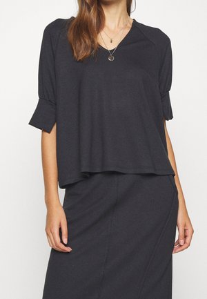 Nainen pukeutuneena löysään tummanharmaaseen yläosaan, jossa on solmittavat puolihihaiset hihat, ja samaa sarjaa olevaan hameeseen, asusteina kerrokselliset kultaiset riipuskaulakorut.