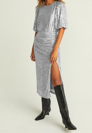 Mujer con vestido midi de lentejuelas plateadas con mangas abullonadas y abertura lateral, combinada con botas negras hasta la rodilla, posando frente a un fondo liso.