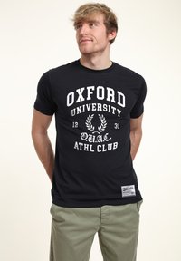Svart kortärmad t-shirt med vit text "Oxford University" och lagerkransgrafik, tillverkad av bomull med en avslappnad passform.