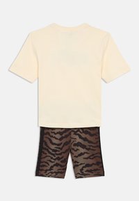 Kremowa koszulka z krótkim rękawem w połączeniu z brązowymi szortami w paski tygrysa, które mają gładką teksturę i czarne akcenty boczne.