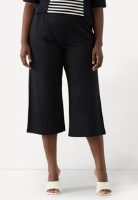 Svarta beskurna byxor med avslappnad passform, elastisk midja och slät textur, parat med öppna vita skor.