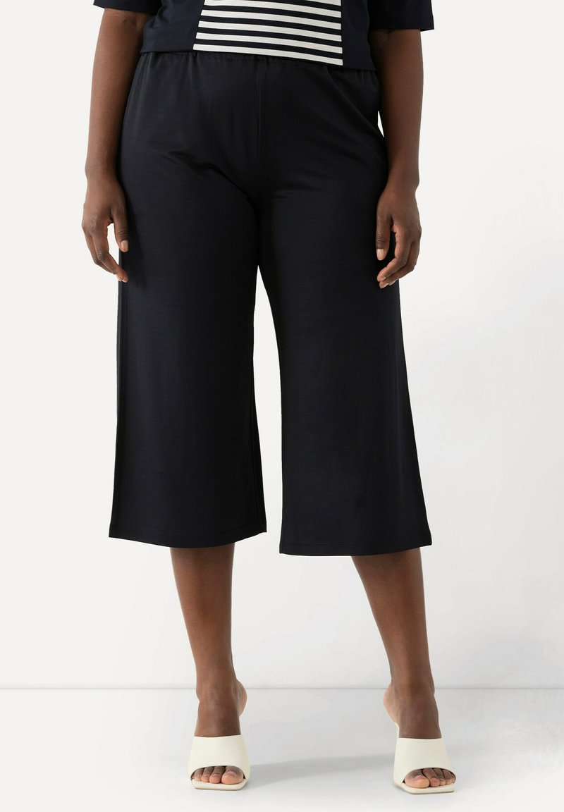 Svarta beskurna byxor med avslappnad passform, elastisk midja och slät textur, parat med öppna vita skor.