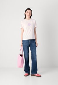 Maglietta di cotone rosa chiaro con logo, jeans svasati blu e borsa tote rosa. Calzature con sneakers rosa con accenti grigi.