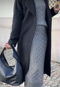 Zwarte wollen jas over een grijze gebreide outfit met een gestructureerd ruitpatroon, met een zwart leren handtas in één hand.