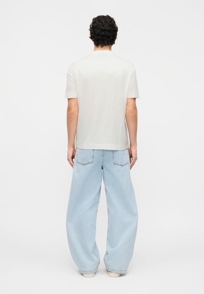 Άνδρας με σγουρά σκουρόχρωμα μαλλιά, φορώντας λευκό κοντομάνικο πουκάμισο και ανοιχτό μπλε τζιν παντελόνι με φαρδιά μπατζάκια, στέκεται με την πλάτη προς τα εμάς σε απλό λευκό φόντο.