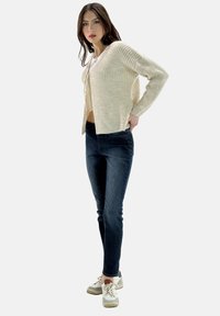 Licht beige gebreid vest met een strikdetail aan de voorkant, gecombineerd met donkerblauwe slim-fit jeans en witte sneakers met grijze accenten.