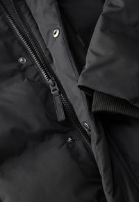 Veste matelassée noire avec fermeture éclair, boutons-pression et poignets côtelés. Le matériau semble lisse avec une finition mate et une texture douce.