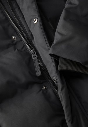 Musta toppatakki, jossa on vetoketjukiinnitys, painonapit ja resorit hihansuissa. Materiaali näyttää sileältä, mattapintaiselta ja pehmeältä.