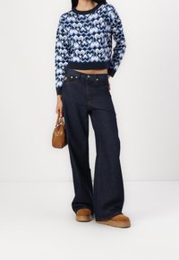 Kobieta ubrana w niebiesko-białą sweter w serca, szerokie ciemne dżinsy, brązowe zamszowe buty i trzymająca małą brązową torebkę.