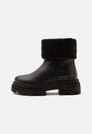 Bottine en cuir noir avec un col texturé épais et une semelle épaisse, avec le logo "Tommy Hilfiger" en or sur le côté.