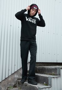 Felpa nera con logo bianco "VANS", pantaloni neri e scarpe slip-on nere. Berretto bordeaux con logo. Modello in piedi su gradini di cemento.