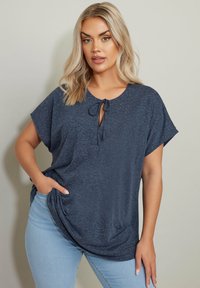 Top blu navy, morbido e a maniche corte, con un dettaglio a fiocco sulla scollatura. Realizzato in un tessuto morbido e testurizzato, abbinato a jeans chiari.