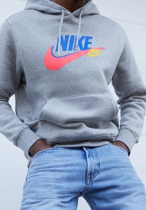 Mężczyzna w szarej bluzie Nike z kolorowymi logo i jasnoniebieskich dżinsach, ręce w kieszeniach, na jednolitym jasnym tle.