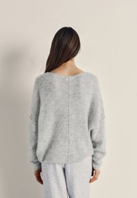 Woman with long brown hair wearing a loose, light gray knit sweater and light gray pants, standing with her back to the camera.