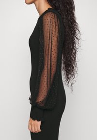 Robe noire côtelée avec des manches bouffantes transparentes à motif pois et des poignets à volants, conçue pour une silhouette ajustée.