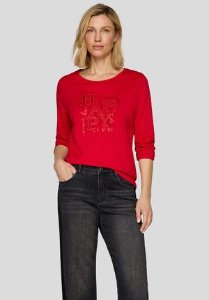 Red long-sleeve t-shirt featuring a graphic print with the word "HAPPY" in bold lettering. Worn with dark denim jeans.