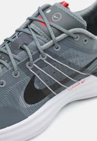 Gros plan sur une chaussure de course Nike grise et noire avec des lacets gris, des accents rouges et une semelle blanche étiquetée "Lunar."