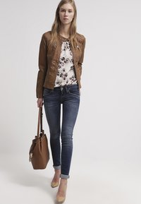 Bruine leren jas over een bloes met bloemen, gecombineerd met donkerblauwe skinny jeans en beige hakken. Een bruin handtas wordt in één hand gedragen.